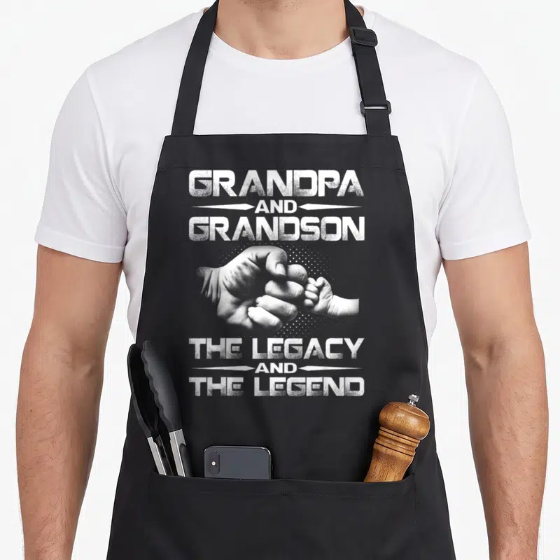 Legend grandpa apron - Distressed white text Grandpa Grandson Legacy Legend above and below clasped adult and baby — shown on a black cotton canvas apron against a white background