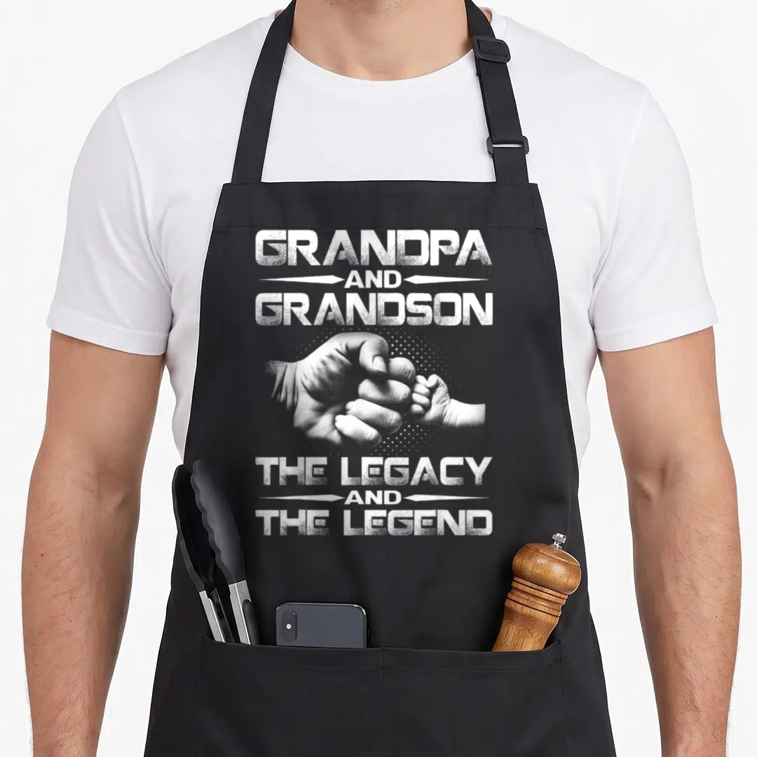 Legend grandpa apron - Distressed white text Grandpa Grandson Legacy Legend above and below clasped adult and baby — shown on a black cotton canvas apron against a white background
