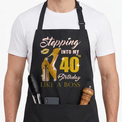 40th bbq boss apron - Gold glitter high heels and pink text reading Stepping Into My 40th Birthday Like A Boss — shown on a black cotton canvas apron against a white background