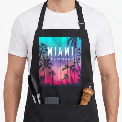 Miami sunset florida apron - Retro palm tree silhouettes over a cyan and pink gradient with centered white MIAMI — shown on a black cotton canvas apron against a white background