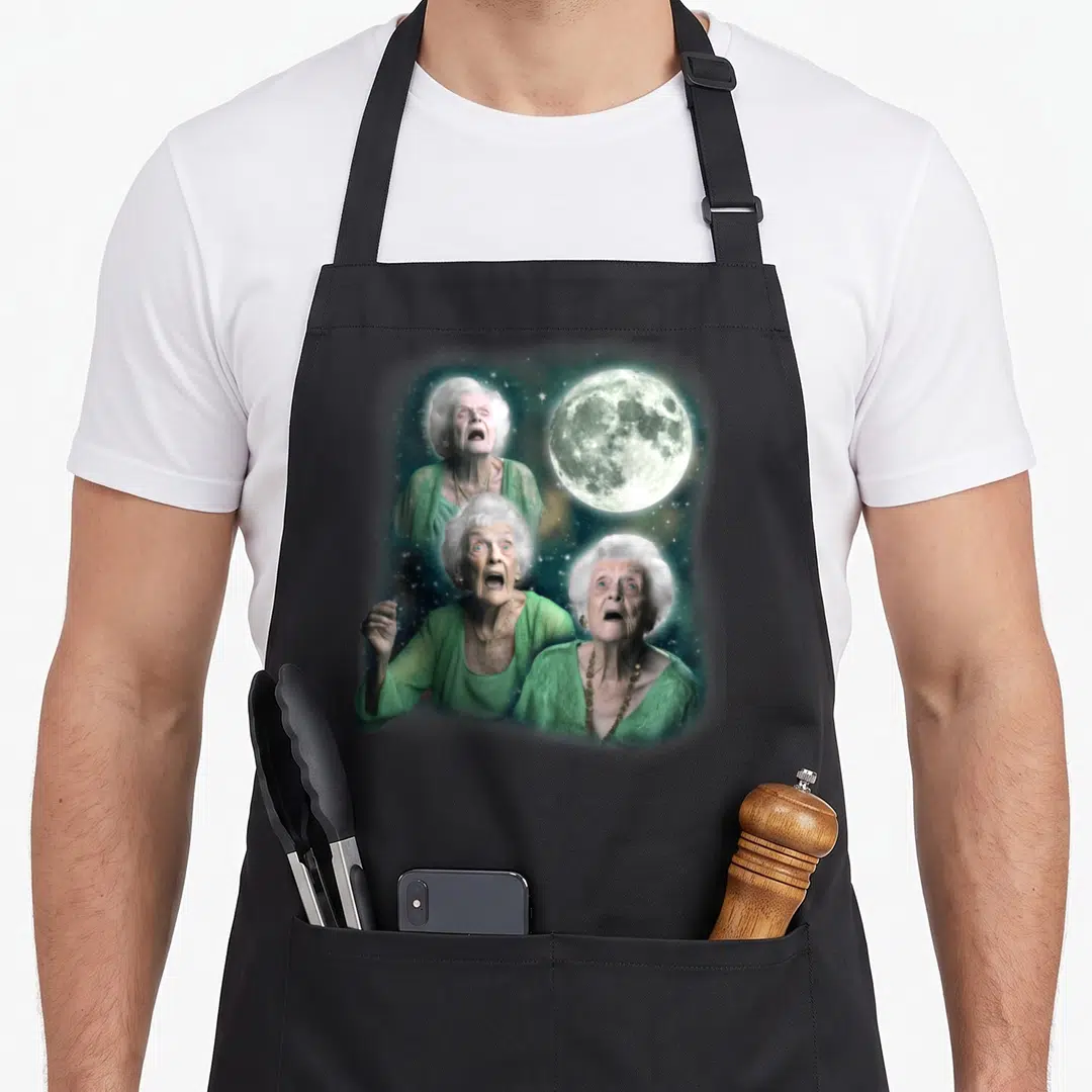 Three granny apron - Three elderly women in green cardsigans posing near a large moon against a starry background — shown on a black cotton canvas apron against a white background