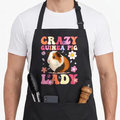 Vintage groovy crazy guinea lady apron - Cartoon guinea pig with CRAZY GUINEA PIG LADY text and colorful flowers — shown on a black cotton canvas apron against a white background