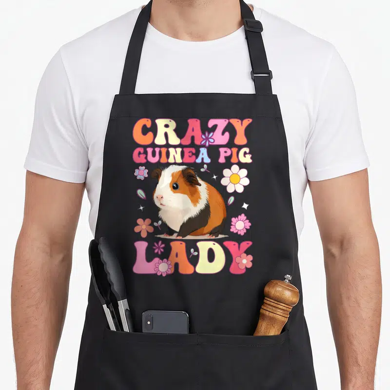 Vintage groovy crazy guinea lady apron - Cartoon guinea pig with CRAZY GUINEA PIG LADY text and colorful flowers — shown on a black cotton canvas apron against a white background