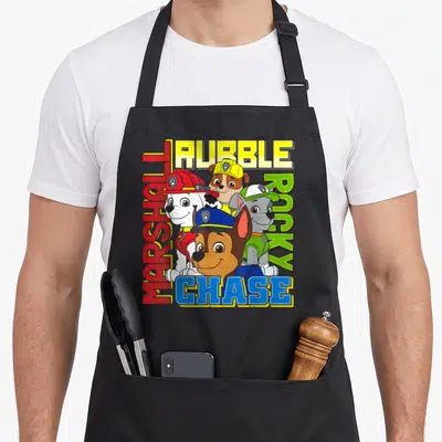 Rocky marshalls apron - Four cartoon dogs surrounded by MARSHALL, RUBBLE, ROCKY, and CHASE text in red, yellow, green — shown on a black cotton canvas apron against a white background
