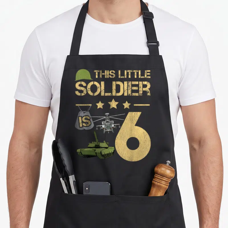 Soldier camo apron - Yellow text THIS LITTLE SOLDIER IS 6 with helicopter tank stars and green helmet on grey background — shown on a black cotton canvas apron against a white background