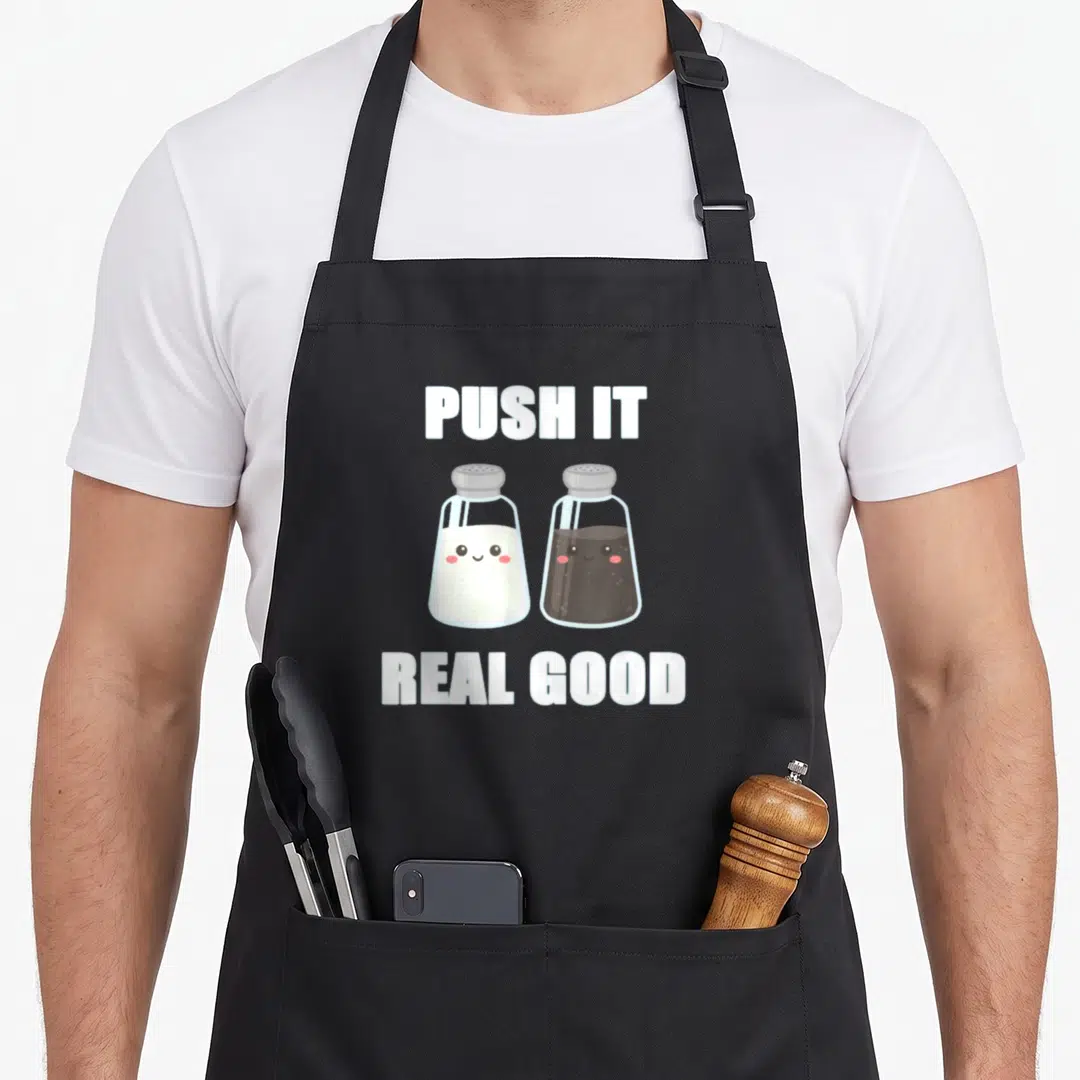 Push salt and pepper aprons - PUSH IT REAL GOOD text above two smiling cartoon salt and pepper shakers in white grey — shown on a black cotton canvas apron against a white background