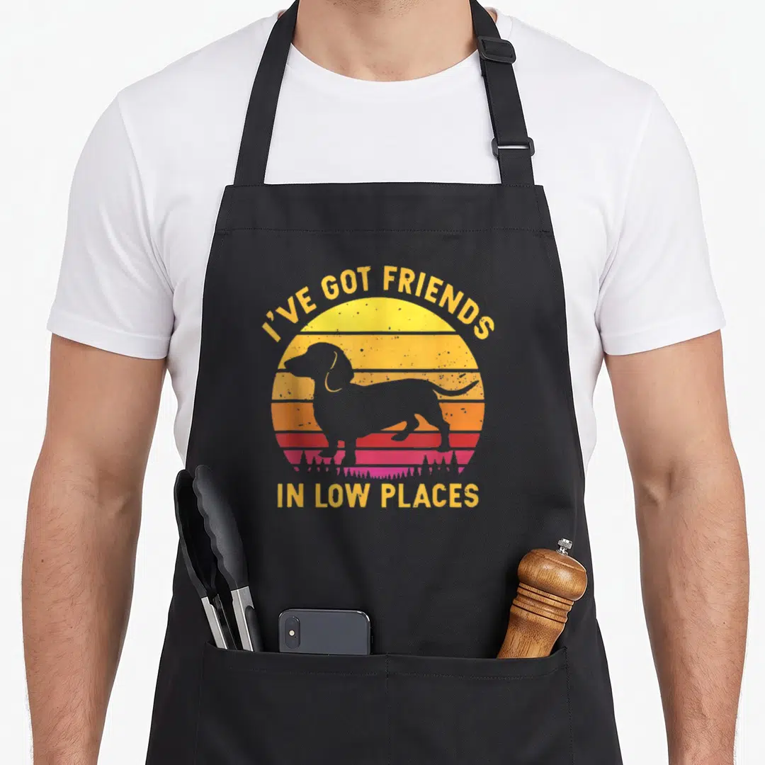 Funny friend dachshund apron - Grey dachshund silhouette over retro striped yellow orange red pink sun with text — shown on a black cotton canvas apron against a white background