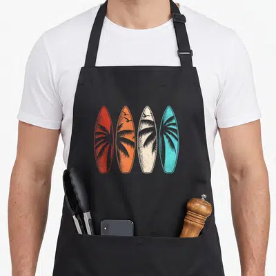 Palm hawaiian apron - Four surfboards in red, orange, cream, and blue with palm trees and birds — shown on a black cotton canvas apron against a white background