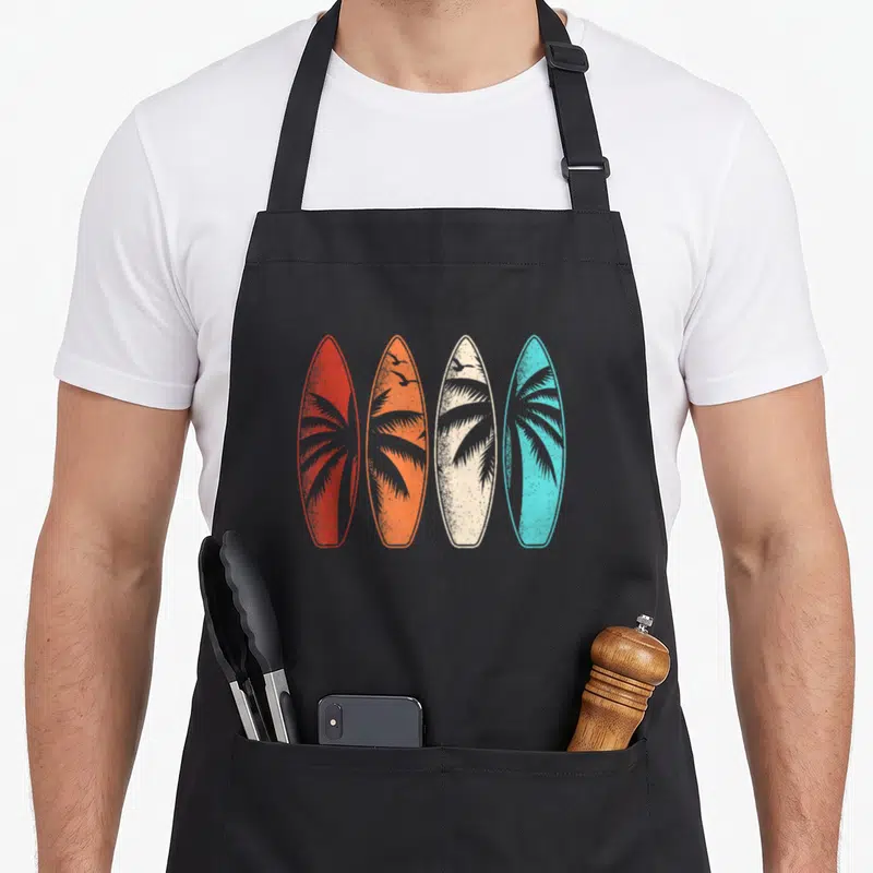 Palm hawaiian apron - Four surfboards in red, orange, cream, and blue with palm trees and birds — shown on a black cotton canvas apron against a white background