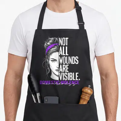 Wound domestic aprons - Woman face with purple bandana and text NOT ALL WOUNDS ARE VISIBLE — shown on a black cotton canvas apron against a white background