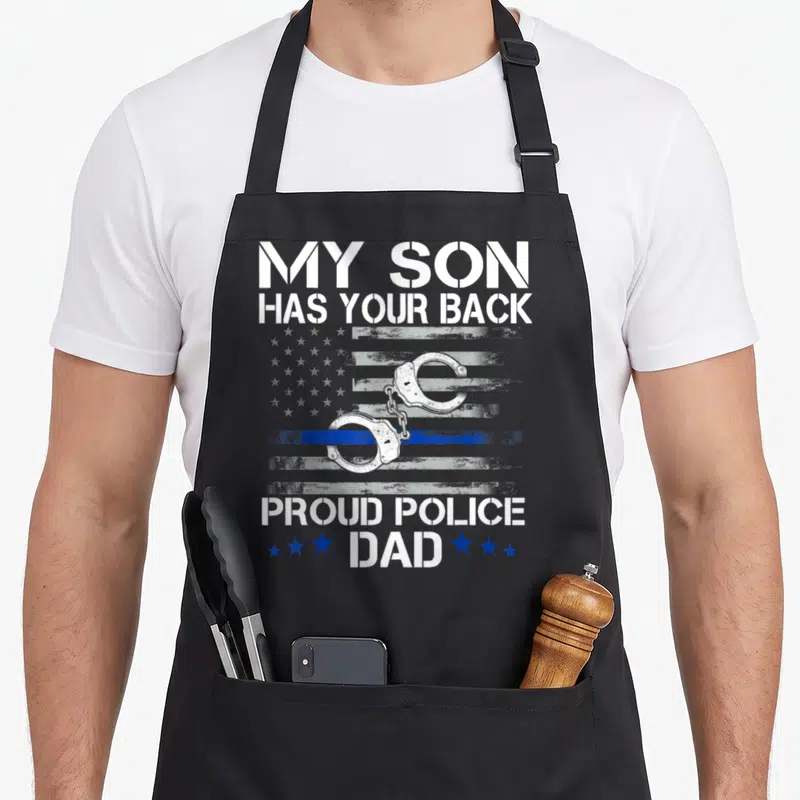 Proud officer thin police apron - My Son Has Your Back text with handcuffs and distressed blue line flag — shown on a black cotton canvas apron against a white background