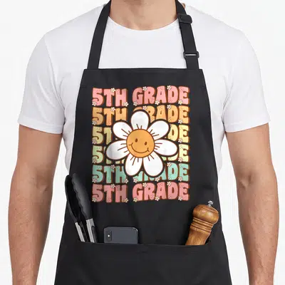 Groovy fifth grade apron - Smiling daisy flower over six stacked lines of 5TH GRADE text in pink orange yellow green — shown on a black cotton canvas apron against a white background