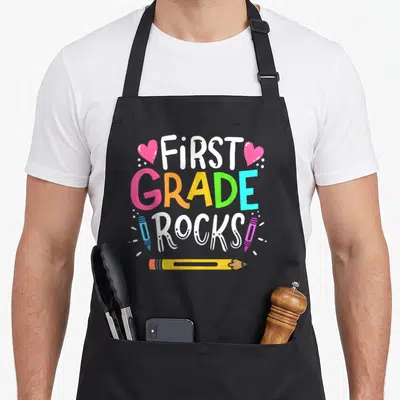 1st art teacher aprons - Stacked FIRST GRADE ROCKS text with pink hearts, blue and pink crayons, yellow pencil, white — shown on a black cotton canvas apron against a white background