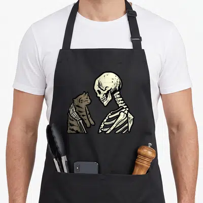Skeleton kitten apron - Cartoon skeleton holding brown cat with bony arm overlap — shown on a black cotton canvas apron against a white background