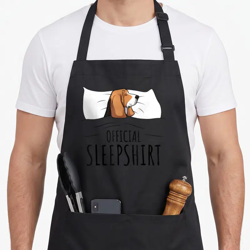 Basset novelty apron - Sleeping bloodhound on white pillow under grey blanket with official sleepshirt text — shown on a black cotton canvas apron against a white background