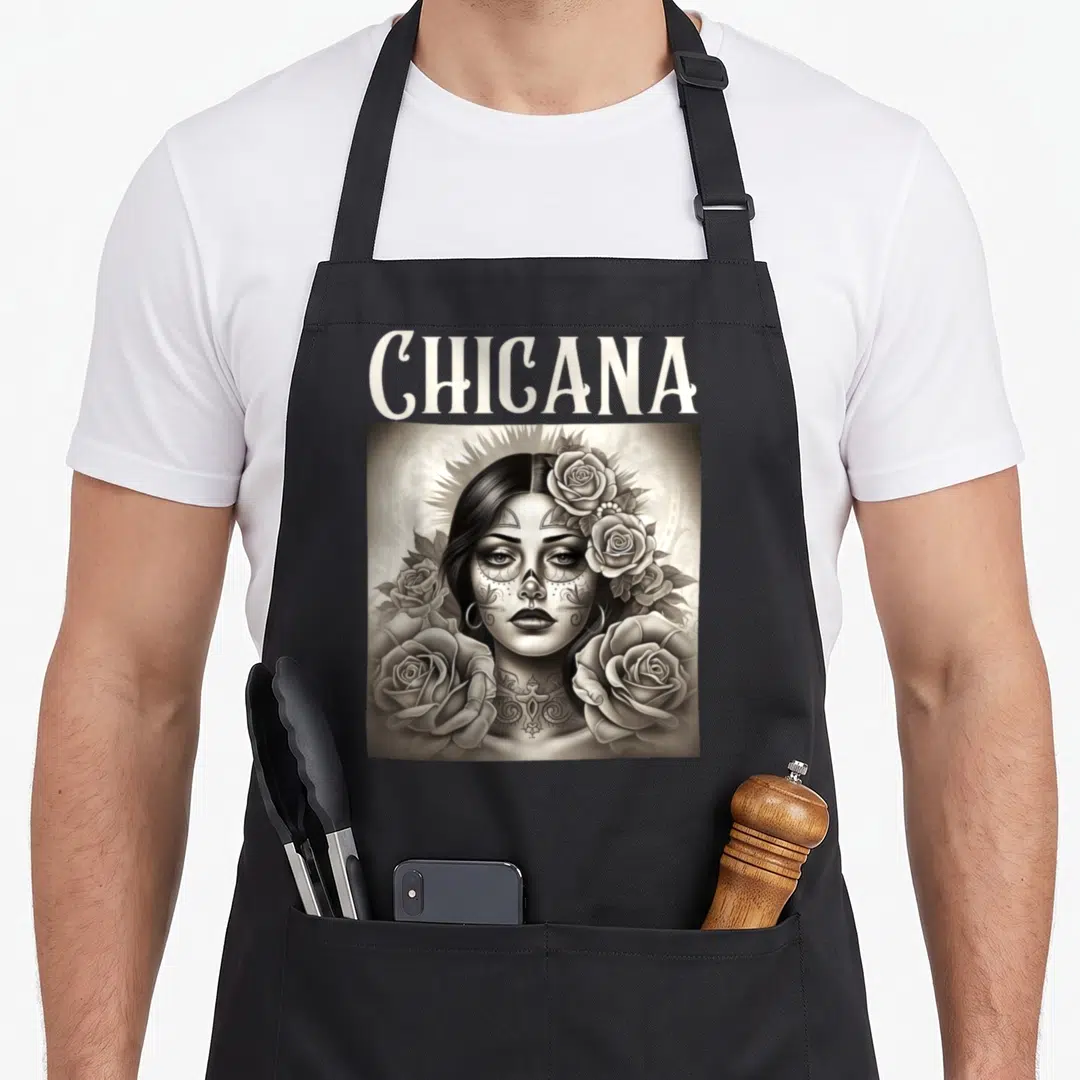 Lowrider mexican style apron - CHICANA text above grayscale woman portrait with sugar skull makeup, tattoos, and roses — shown on a black cotton canvas apron against a white background