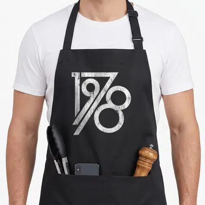 Vintage age birthday apron for grilling - White distressed 1978 typography on black background — shown on a black cotton canvas apron against a white background