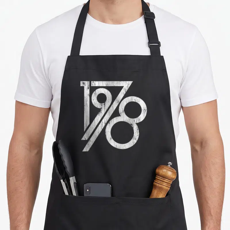 Vintage age birthday apron for grilling - White distressed 1978 typography on black background — shown on a black cotton canvas apron against a white background