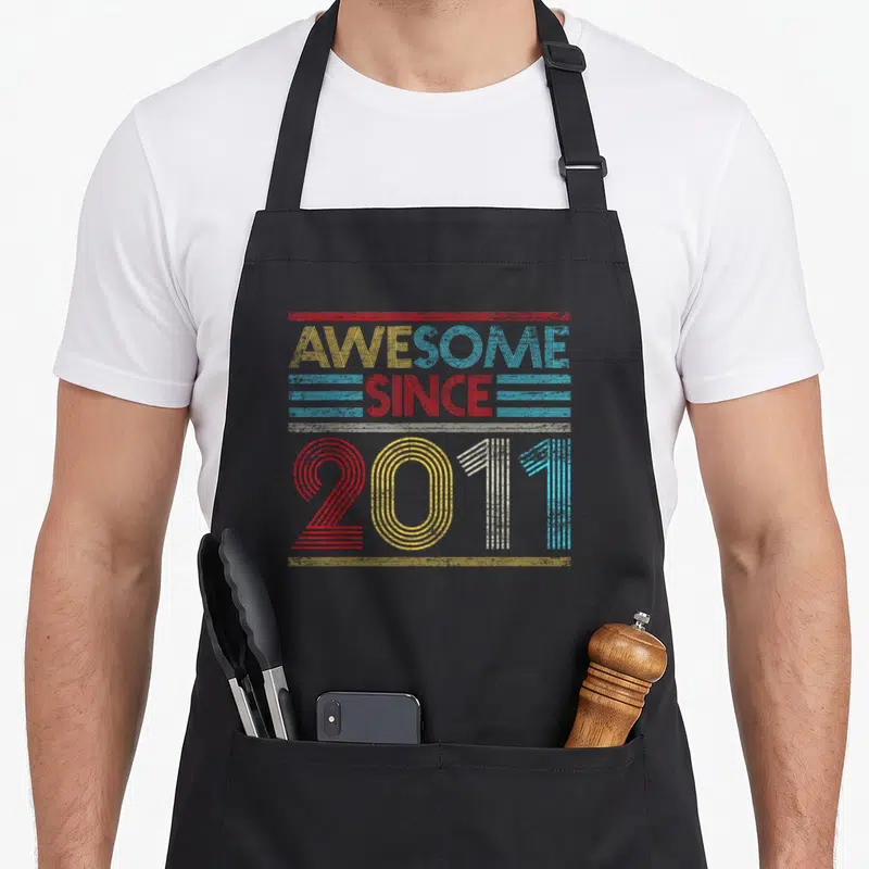 Funny 12th birthday apron - Retro AWESOME SINCE 2011 text design with colorful horizontal stripes — shown on a black cotton canvas apron against a white background