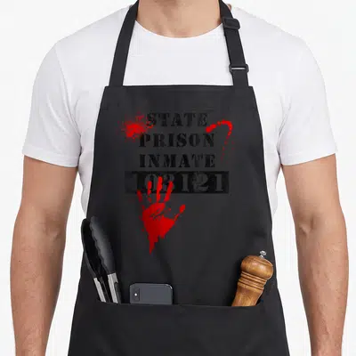 State bloody apron - STATE PRISON INMATE 103121 text and red handprint on black fabric — shown on a black cotton canvas apron against a white background