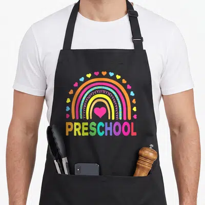 School preschool teacher apron - Rainbow arching over colorful PRESCHOOL text with pink heart center and surrounding — shown on a black cotton canvas apron against a white background