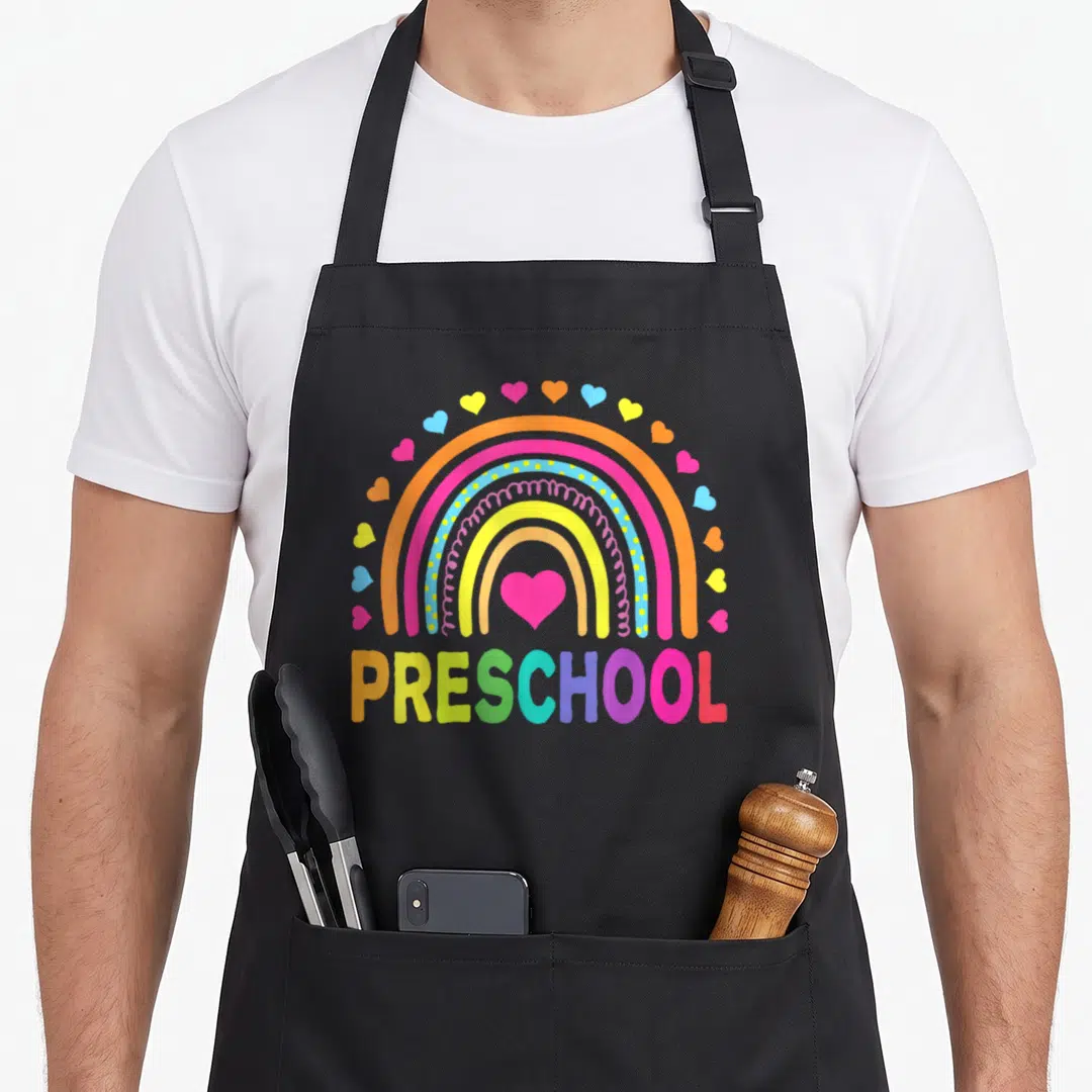 School preschool teacher apron - Rainbow arching over colorful PRESCHOOL text with pink heart center and surrounding — shown on a black cotton canvas apron against a white background
