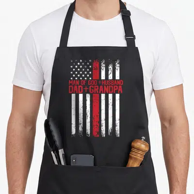 Cross god american flag apron - Distressed US flag with Man of God Husband Dad Grandpa text in red block letters — shown on a black cotton canvas apron against a white background