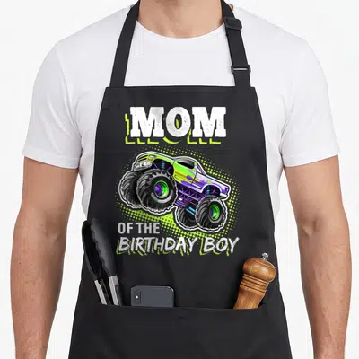 Cute birthday monster truck apron - Monster truck in lime green and purple between MOM and OF THE BIRTHDAY BOY text — shown on a black cotton canvas apron against a white background