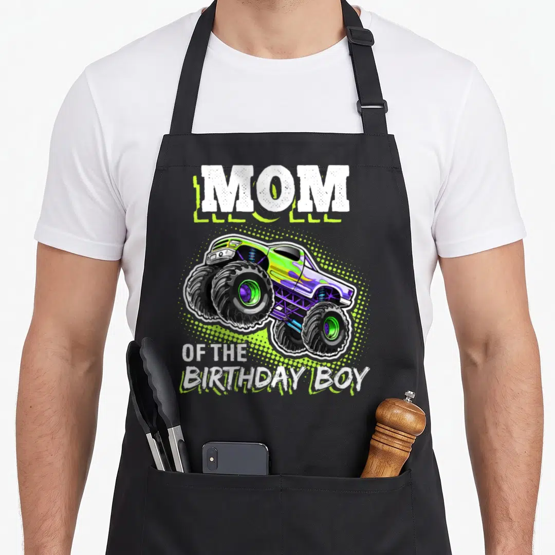 Cute birthday monster truck apron - Monster truck in lime green and purple between MOM and OF THE BIRTHDAY BOY text — shown on a black cotton canvas apron against a white background