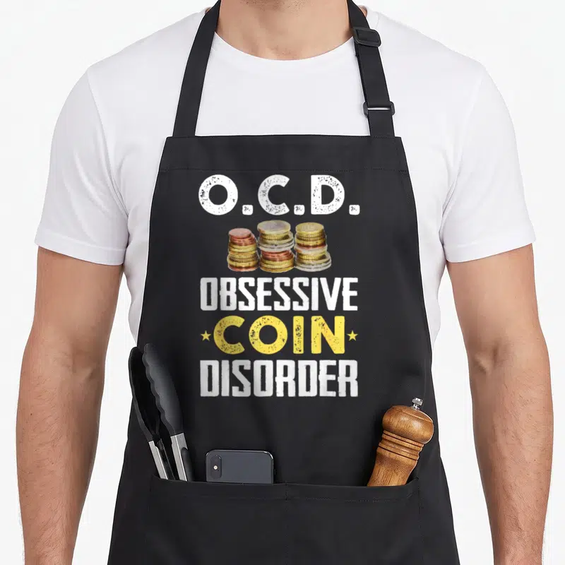 Funny collector coin apron - OCD text above and below three stacks of gold and copper coins with two stars — shown on a black cotton canvas apron against a white background