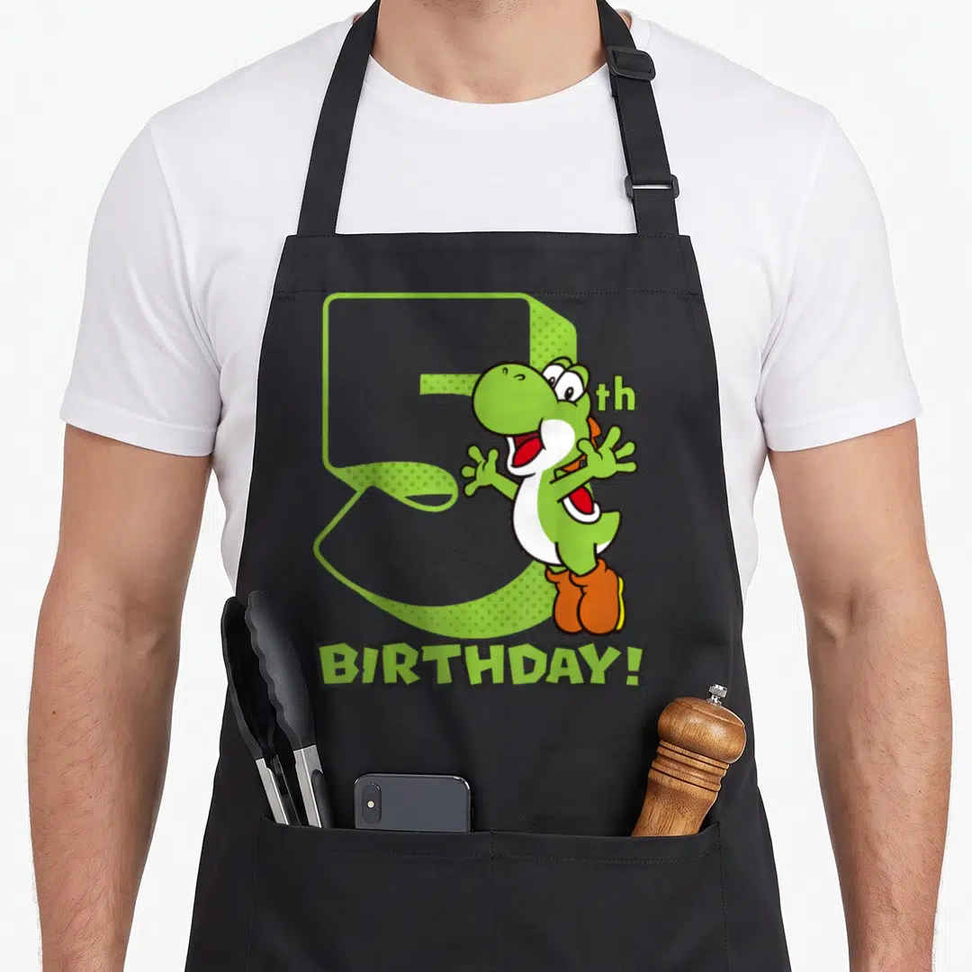 5th holiday apron - Green cartoon dinosaur next to large number 5 with BIRTHDAY text — shown on a black cotton canvas apron against a white background