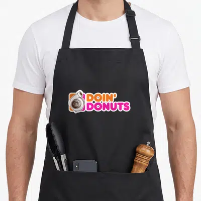 Doin donut car apron - Cartoon turbocharger next to orange and pink DOIN DONUTS text inside white border — shown on a black cotton canvas apron against a white background