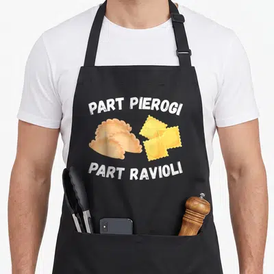 Polish italian aprons - Cartoon pierogi and ravioli with arched text Part Pierogi Part Ravioli in white tan and yellow — shown on a black cotton canvas apron against a white background