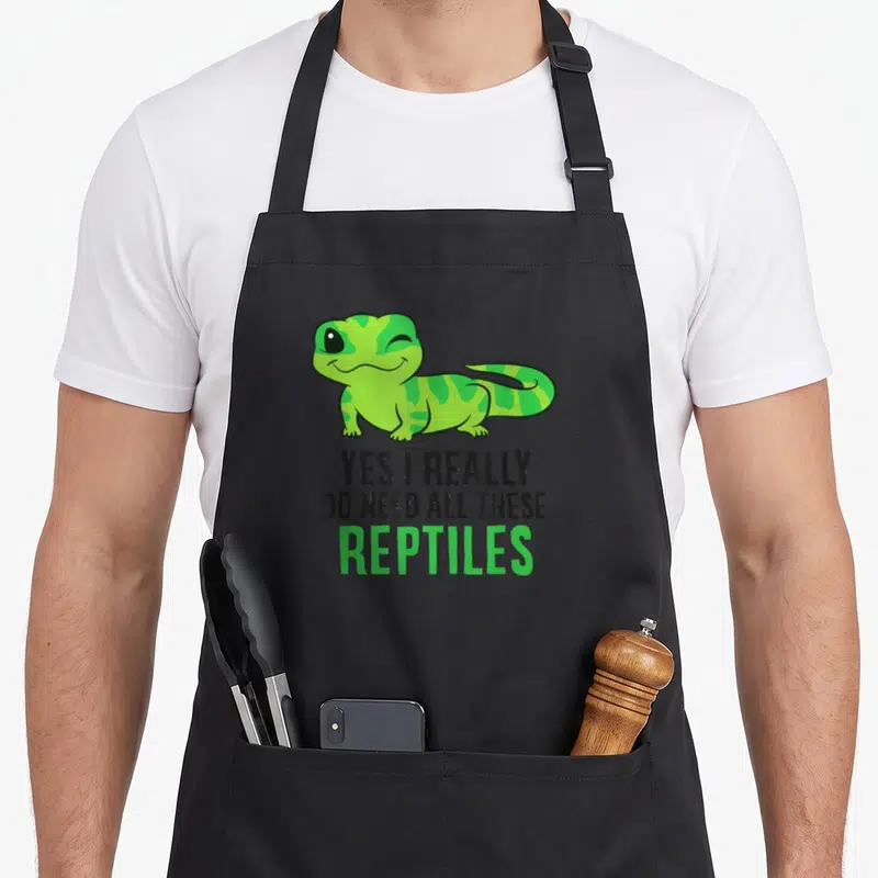 Snake do i need an apron - Green cartoon gecko winking above stacked text reading YES I REALLY DO NEED ALL THESE — shown on a black cotton canvas apron against a white background