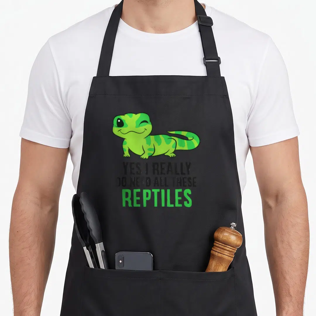 Snake do i need an apron - Green cartoon gecko winking above stacked text reading YES I REALLY DO NEED ALL THESE — shown on a black cotton canvas apron against a white background
