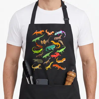Lizard gecko apron - Cartoon lizards in orange, green, blue, red, and yellow scattered on black fabric — shown on a black cotton canvas apron against a white background