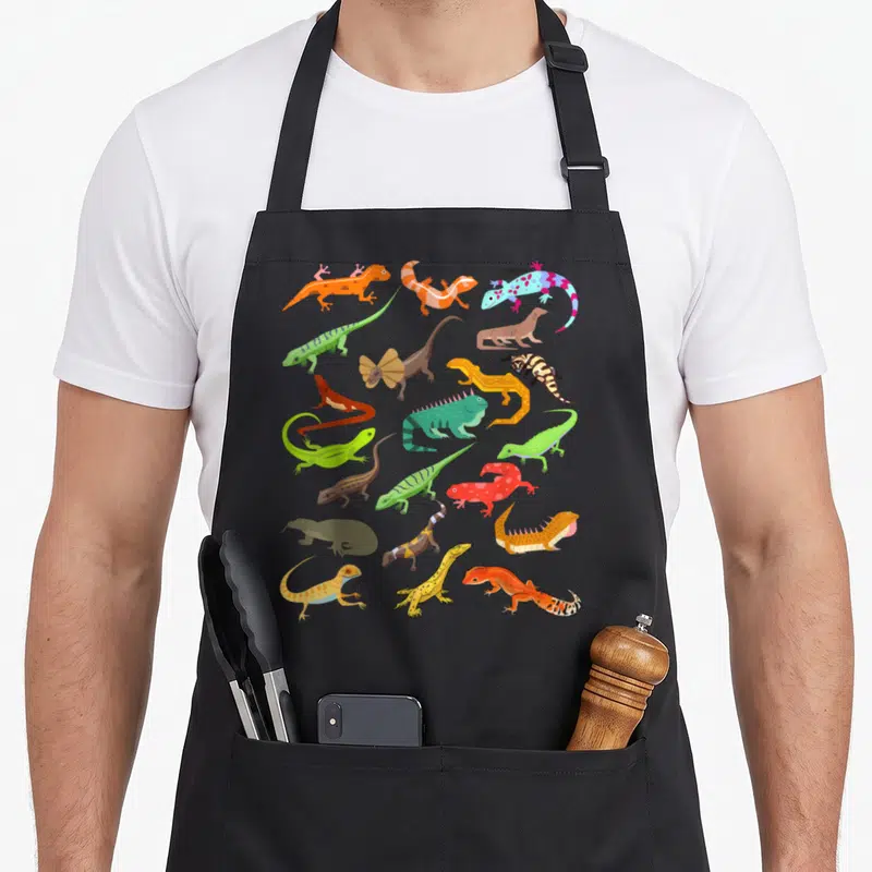 Lizard gecko apron - Cartoon lizards in orange, green, blue, red, and yellow scattered on black fabric — shown on a black cotton canvas apron against a white background