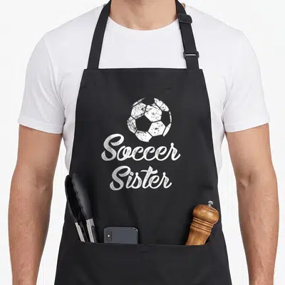 Sister soccer fan apron - Distressed white soccer ball above stacked cursive Soccer Sister text on black fabric — shown on a black cotton canvas apron against a white background