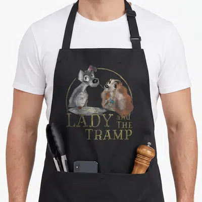 Eating lady and the tramp apron - Two cartoon dogs eating spaghetti from a plate inside a gold circle with stacked text — shown on a black cotton canvas apron against a white background