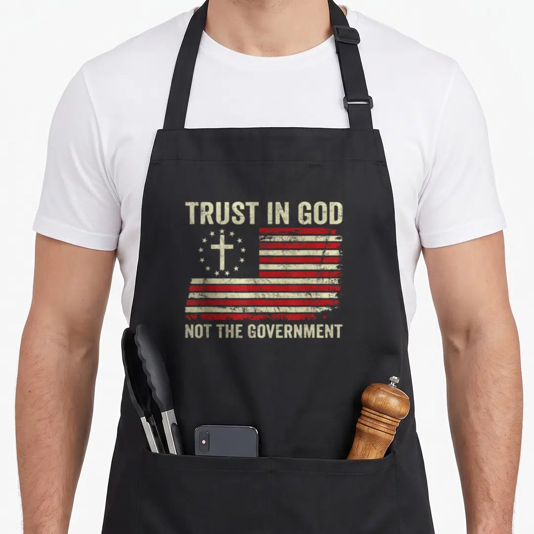 Trust patriotic apron - Distressed flag with cross and TRUST IN GOD NOT THE GOVERNMENT text — shown on a black cotton canvas apron against a white background
