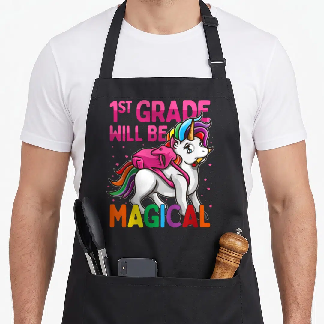 Grade magical unicorn apron - Cartoon unicorn with pink backpack and yellow pencil under 1ST GRADE WILL BE MAGICAL text — shown on a black cotton canvas apron against a white background