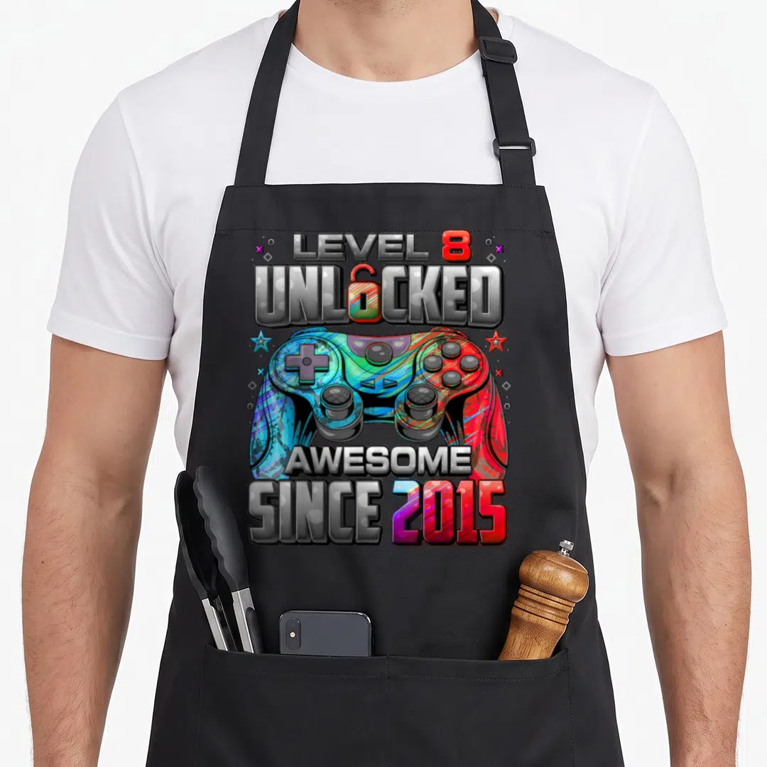 8th gaming aprons - Colorful controller with LEVEL 8 UNLOCKED AWESOME SINCE 2015 text and star icons — shown on a black cotton canvas apron against a white background