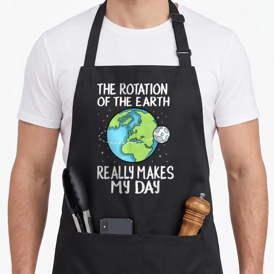 Rotation science apron - Cartoon Earth and moon with dashed orbit line and white pun text — shown on a black cotton canvas apron against a white background