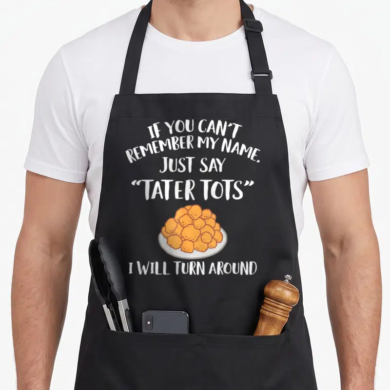 Tot tater apron - Cartoon plate of orange tater tots with white punny text stacked above and below — shown on a black cotton canvas apron against a white background