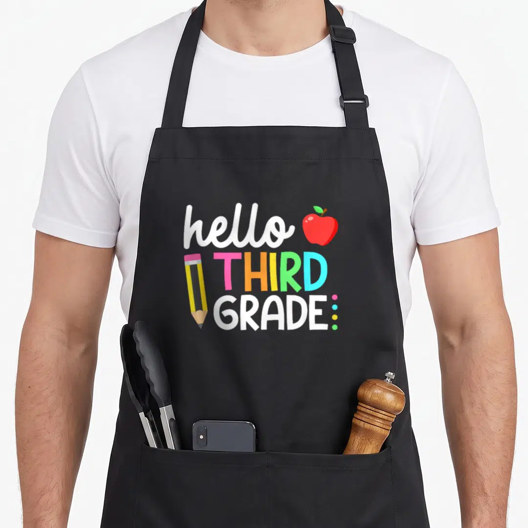 Third teacher apron - Colorful hello THIRD GRADE text with red apple, yellow pencil, and three dots in cartoon style — shown on a black cotton canvas apron against a white background