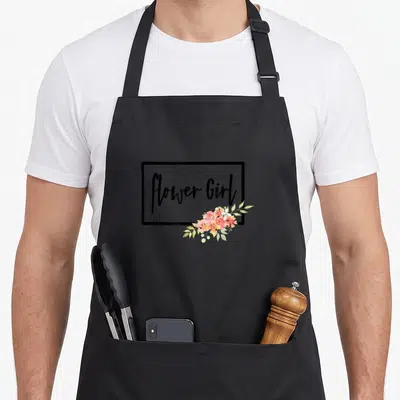 Flower wedding day aprons - Flower Girl text in black frame with pink orange white watercolor flowers at bottom right — shown on a black cotton canvas apron against a white background