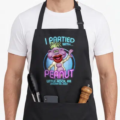 Rock funny peanut apron - Purple monster with I PARTIED WITH PEANUT text and Little Rock date — shown on a black cotton canvas apron against a white background