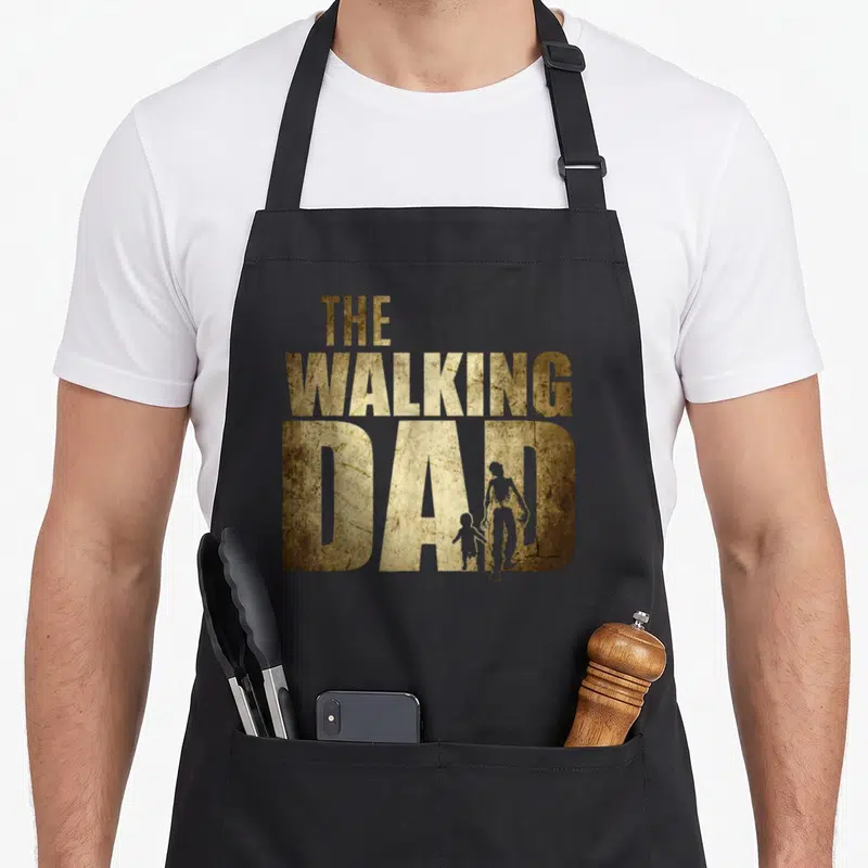 Walking birthday father day apron - Distressed gold text reading THE WALKING DAD with parent and child silhouettes — shown on a black cotton canvas apron against a white background