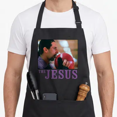 Lebowski bowling apron - Man in purple shirt holding red bowling ball above purple THE JESUS text — shown on a black cotton canvas apron against a white background
