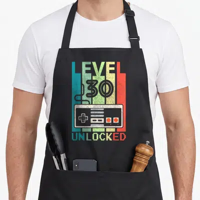 Video gamer 30th birthday apron - Retro game controller with LEVEL 30 UNLOCKED text on four colored vertical stripes — shown on a black cotton canvas apron against a white background